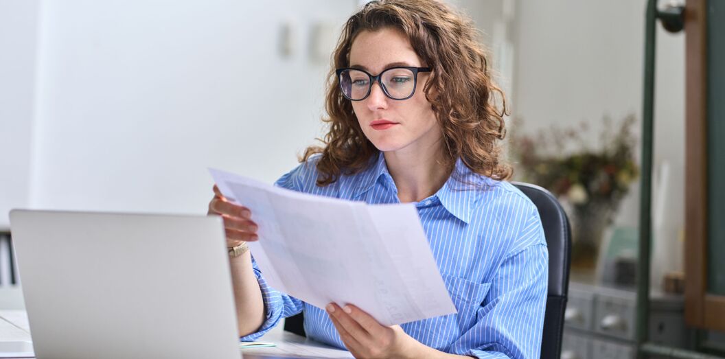 Mitarbeiterin hält Dokumente und arbeitet im Büro mit Laptop.